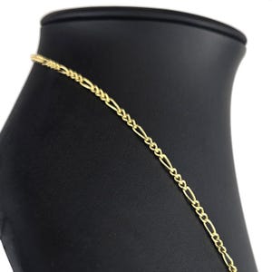 K&ouml;nnte beinhalten: Eine goldfarbene Metallkettenhalskette mit einem klassischen Figaro-Gliedermuster. Die Kette besteht aus einem sich wiederholenden Muster aus drei kleinen runden Gliedern, gefolgt von einem l&auml;nglichen ovalen Glied, was eine gleichm&auml;&szlig;ige Textur ergibt.