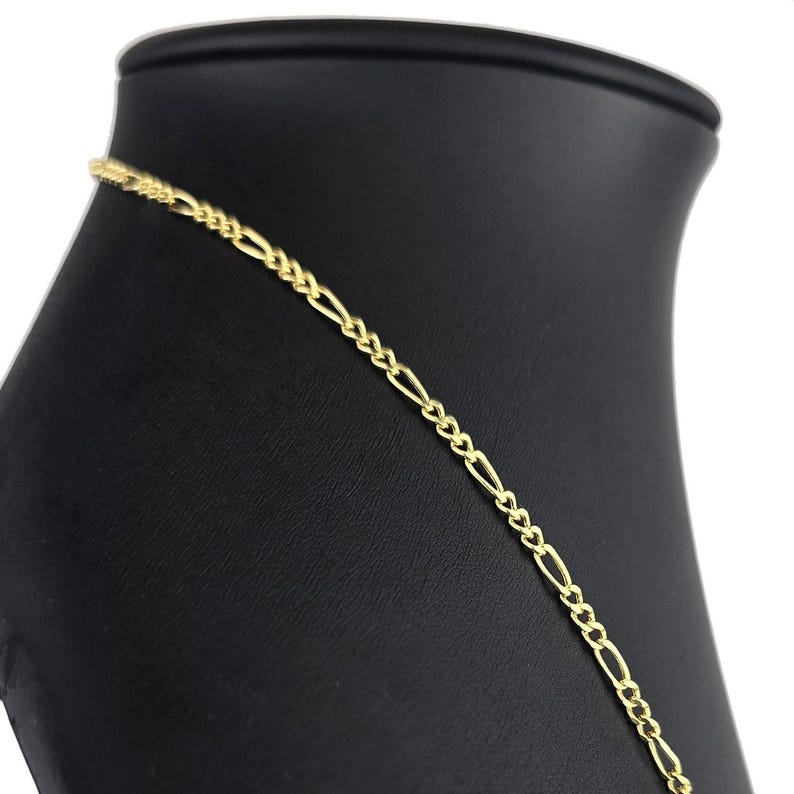K&ouml;nnte beinhalten: Eine goldfarbene Metallkettenhalskette mit einem klassischen Figaro-Gliedermuster. Die Kette besteht aus einem sich wiederholenden Muster aus drei kleinen runden Gliedern, gefolgt von einem l&auml;nglichen ovalen Glied, was eine gleichm&auml;&szlig;ige Textur ergibt.