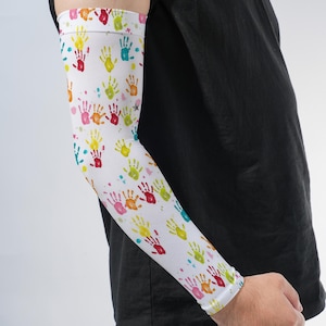 May include: A white arm sleeve featuring a repeating pattern of small, colorful handprints in shades of red, orange, yellow, green, and pink. The sleeve is made of a smooth, stretchy fabric and covers the forearm and elbow area with a snug fit.