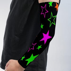 May include: A black arm sleeve featuring a vibrant pattern of neon green, hot pink, and orange stars of varying sizes. The sleeve covers the forearm and extends toward the upper arm, displaying a high-contrast graphic design against the black fabric.
