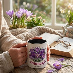 Może przedstawiać: Biały ceramiczny kubek z czarnym wnętrzem i uchwytem, ​​z fioletowym wzorem kwiatu krokusa i tekstem "#FAITHING" i "MATT 11:28-30". Kubek trzymany jest przez osobę, z książką i okularami w tle.
