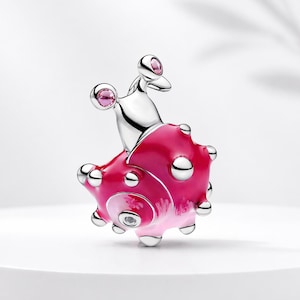 Puede incluir: Un charm de plata con un diseño de caracol rosa y blanco. El caracol tiene una concha texturizada con detalles plateados y gemas rosas. El charm está sobre un fondo blanco.