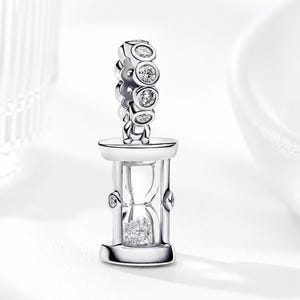 May include: A silver-colored charm in the shape of an hourglass, with clear glass and sparkling accents. The charm has a series of round, clear stones along the top. The hourglass contains small, clear stones. The charm is likely for a bracelet or necklace.