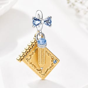 May include: A gold-colored charm with a graduation cap design, a pencil, and a blue bow. The charm has a silver loop and a blue gemstone on the cap. The pencil is gold with a blue gemstone eraser. A small blue gemstone is on the cap.