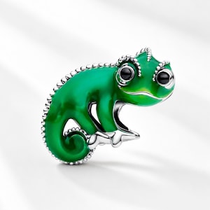 Pode incluir: Um berloque de camaleão verde com acabamento brilhante e olhos pretos. O berloque tem uma borda prateada e é projetado em um estilo detalhado e realista. O camaleão está posicionado em uma postura curvada.