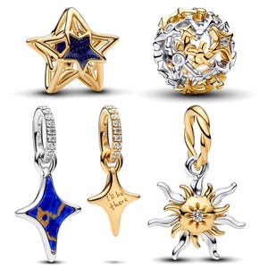 Puede incluir: Una colección de cinco dijes de joyería que incluye una estrella dorada con un centro azul brillante, un dije esférico con recortes de estrellas doradas y plateadas, dos colgantes en forma de estrella con cristales y un dije de sol.