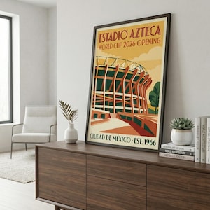 May include: Framed poster featuring the Estadio Azteca stadium, with text reading "ESTADIO AZTECA WORLD CUP 2026 OPENING CIUDAD DE MÉXICO EST. 1966." The artwork depicts the stadium in orange, green, and brown tones. The poster is displayed on a dark wood cabinet.