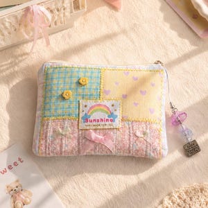 May include: A rectangular fabric pouch featuring a patchwork design of blue plaid, yellow with purple hearts, and pink floral patterns. It includes a central patch with a rainbow and the text Sunshine HAND MADE FOR YOU, plus small bows and flower embellishments.