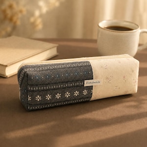 Può includere: Un astuccio rettangolare in tessuto con un design bicolore. Il lato sinistro è grigio scuro con motivi floreali e a righe bianche, mentre il lato destro è color crema con una sottile stampa floreale. È presente una piccola etichetta bianca con la scritta Handmade.