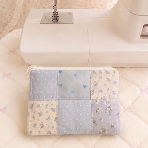 May include: A rectangular zippered pouch featuring a patchwork design of light blue and cream fabric squares. The pattern includes small white polka dots, tiny blue stars, and delicate floral prints in soft blue and cream tones with a white zipper.