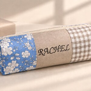 May include: A rectangular pencil case featuring a blue floral print on one end, a beige center panel embroidered with the name RACHEL in black, and a brown and white gingham check pattern on the other end, finished with a top zipper closure.