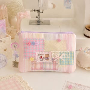 Op de afbeelding: Een rechthoekig stoffen etui met rits en een patchworkontwerp van pastelkleurige ruitpatronen in roze, geel en blauw. Het etui bevat een kleine geborduurde patch van een beer en een cupcake met de tekst "Cake", bloemvormige knopen, kanten randen en een bungelende bedel.