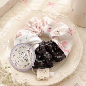 May include: A fabric hair scrunchie featuring a combination of white and pink gingham, white fabric with small bunny and floral prints, and a dark brown section with a floral pattern. A round tag reads Violet Cozy Club A little package of happiness.