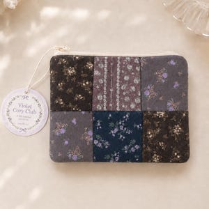 May include: A rectangular fabric zip pouch featuring a patchwork design of six squares with dark floral and butterfly patterns in shades of purple, navy, and black. A round paper tag attached by a string displays the text Violet Cozy Club.