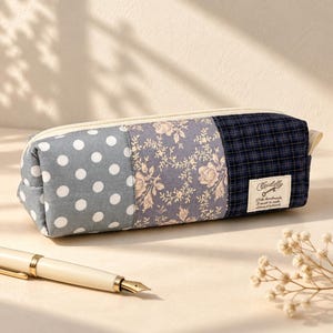 Puede incluir: Un estuche rectangular de tela con un diseño de patchwork de tres secciones: un panel gris con lunares blancos, un panel con estampado floral gris y un panel de cuadros azul oscuro con una etiqueta blanca que dice Cuddly.