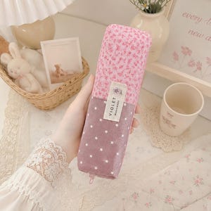 Könnte beinhalten: Ein rechteckiges Federmäppchen mit zweifarbigem Design, bestehend aus einem oberen Abschnitt mit rosa Blumenmuster und einem unteren Abschnitt in Mauve, der mit kleinen weißen Sternen verziert ist. Ein Stoffetikett trägt VIOLET HANDMADE.
