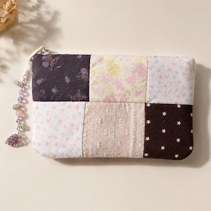 May include: A rectangular patchwork pouch with a zipper closure and a beaded charm. The pouch is made of fabric squares in various patterns, including floral, stars, and lace. The color palette includes purple, pink, white, and brown.