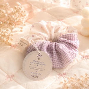 May include: A fabric hair scrunchie featuring a split design with one half in a white floral print and the other half in a light purple gingham check pattern. A round paper tag is attached, printed with the text Thank you for supporting handmade.