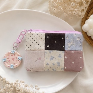 May include: A patchwork coin purse with a light pink zipper and a matching keychain. The purse is made of various fabric squares in cream, pink, and blue, with polka dots, floral, and star patterns. The keychain is round and decorated with blue and pink embellishments.