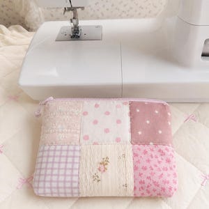May include: A rectangular zippered pouch constructed from a patchwork of various pink and cream fabrics. The design features squares with patterns including light pink polka dots, a lavender gingham check, small white stars, and floral prints.