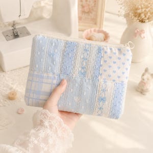 Puede incluir: Un estuche rectangular con cremallera que presenta un diseño de patchwork de telas de algodón en azul claro y blanco. Los patrones incluyen pequeños corazones azules, cuadros y un intrincado bordado floral tipo encaje en cuadrícula.