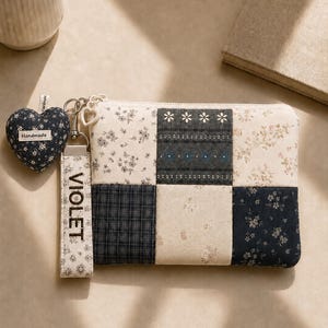 May include: A rectangular zipped pouch featuring a patchwork design of navy blue, cream, and grey fabrics with floral and plaid patterns. A wrist strap with the text VIOLET and a heart-shaped charm with the text Handmade are attached to the zip.
