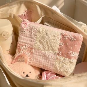 May include: A rectangular zipped pouch featuring a patchwork design of pink gingham, cream lace, and star-patterned fabric. The pouch has a heart-shaped zip pull and is nestled inside a large cream canvas tote bag with a water bottle and plush toy.