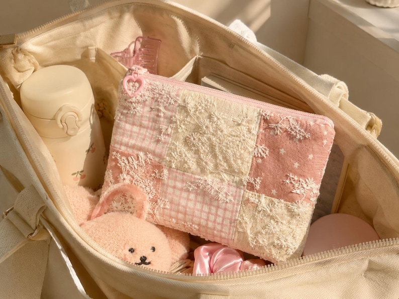 May include: A rectangular zipped pouch featuring a patchwork design of pink gingham, cream lace, and star-patterned fabric. The pouch has a heart-shaped zip pull and is nestled inside a large cream canvas tote bag with a water bottle and plush toy.