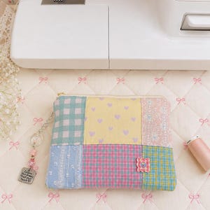 Puede incluir: Un estuche rectangular de patchwork con cremallera que presenta paneles de tela amarilla con corazones morados, cuadros vichy azules y blancos, estampado floral y cuadros rosas. Un dije plateado en la cremallera dice "Follow your dreams Trust your heart."