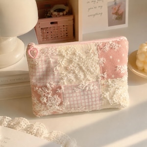 May include: A rectangular zipped pouch featuring a patchwork design of pink gingham, solid pink fabric with white stars, and cream-coloured floral lace overlays. The zip pull is a small, light pink plastic heart-shaped charm.