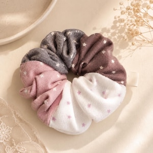 May include: A circular fabric hair scrunchie featuring four distinct segments in shades of charcoal gray, muted brown, dusty pink, and white. Each segment is patterned with small, evenly spaced star shapes in contrasting colors.
