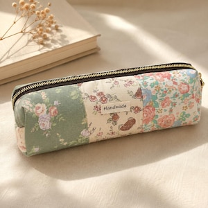 May include: A rectangular fabric pencil case featuring a patchwork design of floral patterns in shades of green, cream, and pink. The case has a dark brown zip closure and a small rectangular label with the word Handmade stitched on the front.