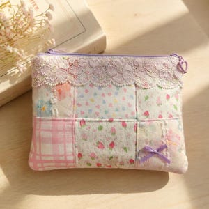 Peut inclure: Une pochette rectangulaire à fermeture éclair avec un motif patchwork de divers tissus pastel, incluant des carreaux roses, des imprimés fraises et des motifs floraux. Le bord supérieur est orné de dentelle lavande et d'un petit nœud.