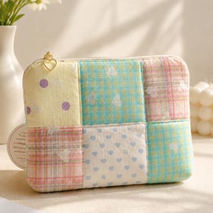 May include: A rectangular fabric pouch featuring a patchwork design of pastel yellow, teal, and pink squares with patterns including polka dots, plaid, and small hearts. The pouch has a gold-toned zip with a heart-shaped pull tab.