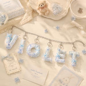 May include: A decorative garland featuring plush, fabric-covered letters spelling VIOLET in a light blue and white gingham pattern. Each letter is adorned with a small blue ribbon and attached to a silver-toned metal chain with jump rings.