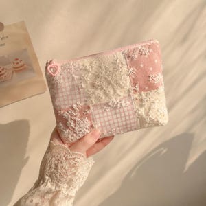 May include: A rectangular zipped pouch featuring a patchwork design of light pink gingham, solid pink fabric with white stars, and cream-coloured lace floral overlays. The zip pull is a small, light pink heart-shaped plastic charm.