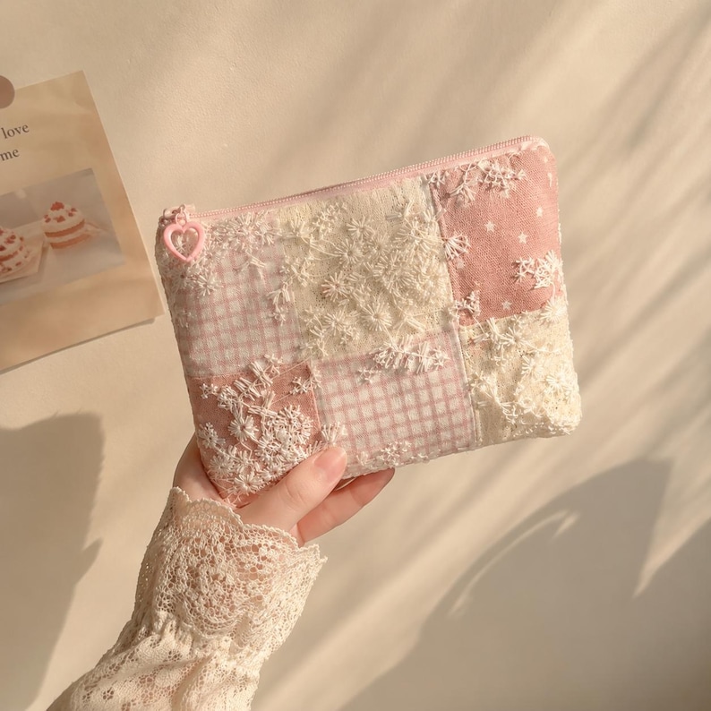 May include: A rectangular zipped pouch featuring a patchwork design of light pink gingham, solid pink fabric with white stars, and cream-coloured lace floral overlays. The zip pull is a small, light pink heart-shaped plastic charm.