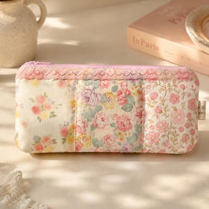 May include: A rectangular fabric pencil case featuring a patchwork design of various floral patterns in shades of pink, yellow, and green. The case has a pink zipper closure topped with a delicate lace trim and a small side tag with text.
