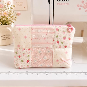 May include: A rectangular fabric pouch featuring a patchwork design of pink and cream floral, strawberry, and polka dot patterns. The pouch includes white lace trim, a small pink ribbon bow, and a pink heart-shaped zipper pull.
