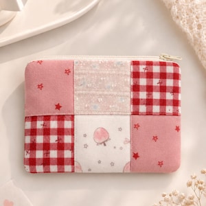 Könnte beinhalten: Eine rechteckige Patchwork-Geldbörse mit Reißverschluss. Die Geldbörse hat eine Mischung aus rosa Stoffen, darunter ein Vichy-Muster, Stoff mit Sternenaufdruck und Stoff mit einer Pfirsich-Illustration. Der Reißverschluss ist goldfarben.