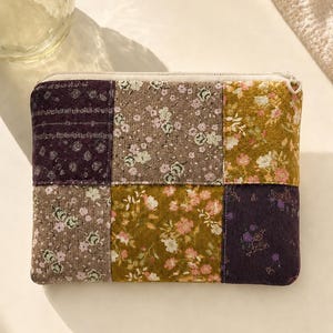 Könnte beinhalten: Eine rechteckige Reißverschlusstasche mit einem Patchwork-Design aus sechs Stoffquadraten. Die Stoffmuster umfassen dunkelviolette, gedeckte braune und senfgelbe Blumenprints mit kleinen weißen, rosafarbenen und grünen Blumendetails.