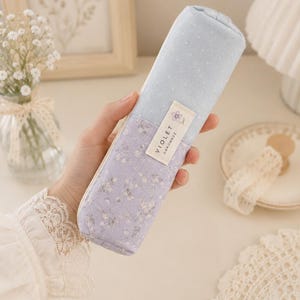 Peut inclure: Une trousse à crayons rectangulaire en tissu au design bicolore, avec une partie supérieure bleu clair à pois blancs et une partie inférieure lavande à motif floral. Une étiquette en tissu blanc porte l'inscription VIOLET HANDMADE.