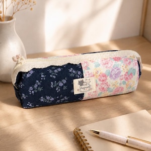 May include: A rectangular fabric pencil case featuring a patchwork design of dark blue floral fabric and a light pink and yellow floral print. A cream-colored lace trim borders the zipper, and a small label reads Handmade with a cat illustration.