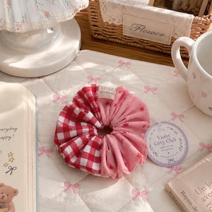 May include: A fabric hair scrunchie featuring a split design with one half in a red and white gingham check pattern and the other half in a solid pink fabric with small red star prints. A small white tag reads VIOLET HANDMADE.