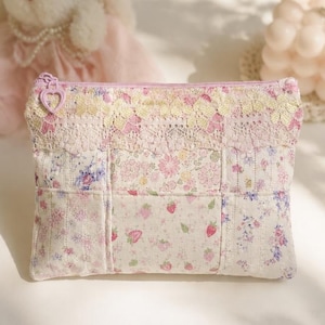 Könnte beinhalten: Ein rechteckiges Stofftäschchen mit einem Patchwork-Design aus Blumen- und Erdbeermustern in Rosa-, Weiß- und Blautönen. Die Oberkante ist mit einem cremefarbenen Spitzenband und einem herzförmigen Reißverschlussanhänger verziert.