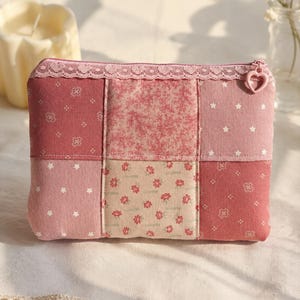 May include: A rectangular zippered pouch featuring a patchwork design of various pink and red fabrics with floral, star, and geometric patterns. The top edge is trimmed with a delicate pink lace border and a small heart-shaped zipper pull.