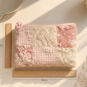 May include: A rectangular zippered pouch featuring a patchwork design of pink gingham, solid pink fabric with white stars, and cream-coloured floral lace overlays. The pouch includes a heart-shaped zipper pull and measures 12 cm by 16 cm.