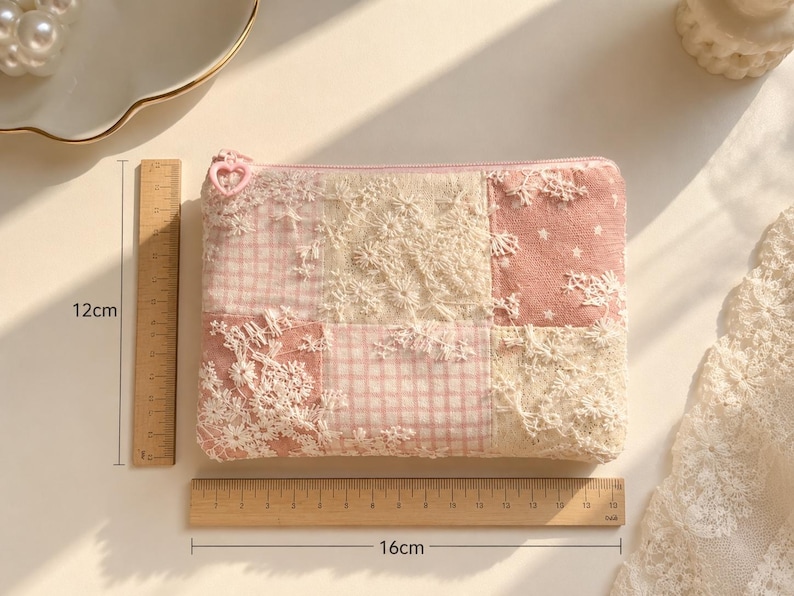 May include: A rectangular zippered pouch featuring a patchwork design of pink gingham, solid pink fabric with white stars, and cream-coloured floral lace overlays. The pouch includes a heart-shaped zipper pull and measures 12 cm by 16 cm.