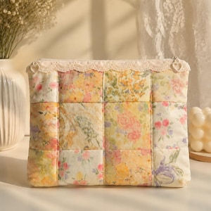 Puede incluir: Un estuche rectangular con cremallera que presenta un diseño de patchwork con varios tejidos florales en tonos amarillos, rosas y verdes. El borde superior está rematado con un encaje color crema y un tirador de metal en forma de corazón.