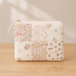 Könnte beinhalten: Eine rechteckige Reißverschlusstasche mit gestepptem Patchwork-Design. Der Stoff zeigt verschiedene florale Muster in Rosa-, Gelb-, Blau- und Grüntönen auf cremefarbenem Grund. Ein dünner Stoffanhänger ist am oberen Reißverschluss befestigt.